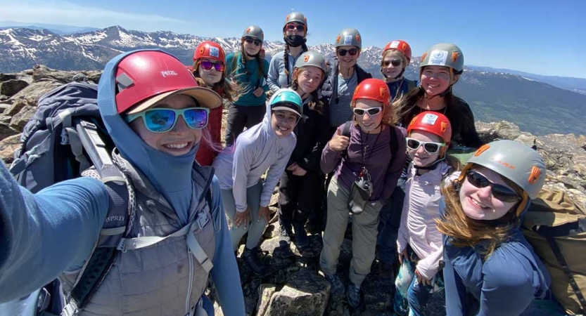 A group of people wearing helmets stand at high elevation over a mountain landscape and smile for the photo. 
